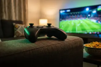 Black wireless game controller on a sofa armrest with a football match on a TV behind