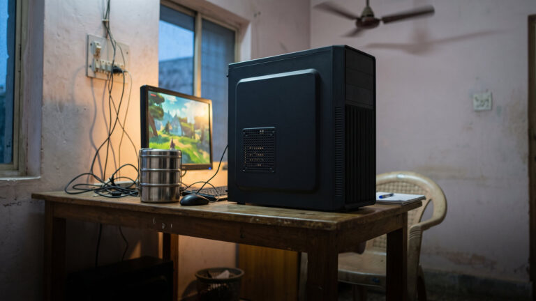 Plain black budget PC cabinet with no lights on a simple wooden table beside a monitor