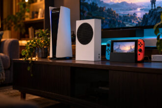 Three unbranded game consoles, a white tower, a small white box and a handheld in a dock, on a living room shelf