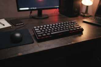 Compact black 60 percent keyboard with a red backlight beside a wired mouse on a student desk