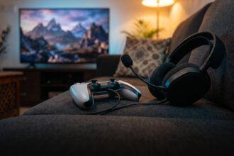 Gaming headset plugged into a white game controller on a sofa with a TV glowing behind