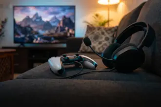 Gaming headset plugged into a white game controller on a sofa with a TV glowing behind