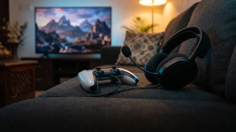 Gaming headset plugged into a white game controller on a sofa with a TV glowing behind