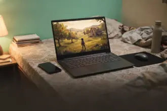 Thin budget laptop on a floor mattress showing a hillside orchard game, phone hotspot beside it
