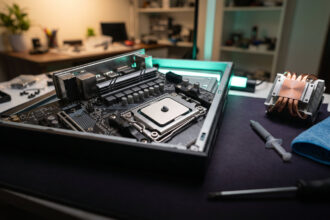 Pea sized dot of grey thermal paste on a CPU heatspreader with a syringe and cooler nearby on a workbench