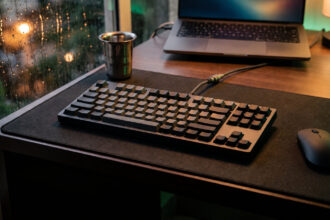 Dark grey tenkeyless keyboard with arrow keys and a mouse on a desk beside a rainy window