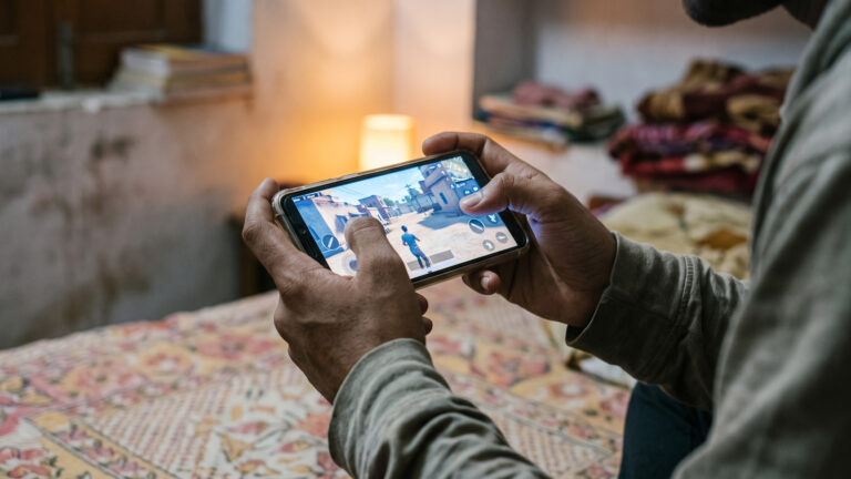 Hands holding a budget Android phone sideways, playing a mobile shooter on a bed at night