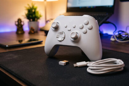 Slim white wireless controller with offset sticks on a desk beside a small USB dongle, coiled cable and a phone
