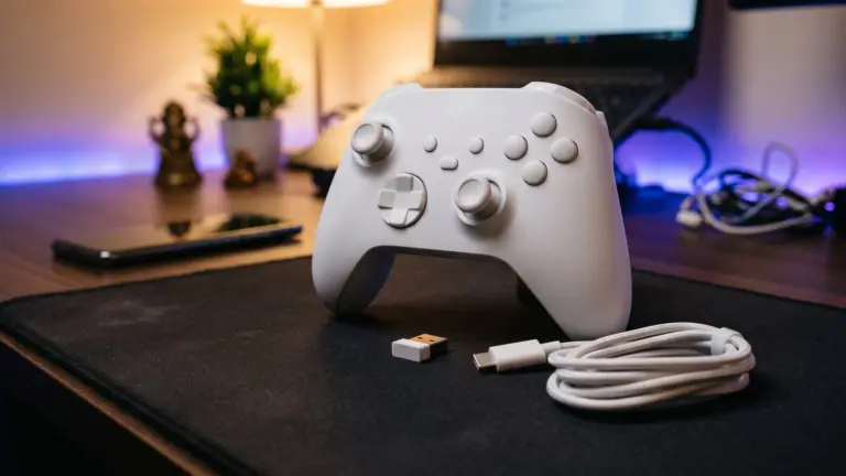 Slim white wireless controller with offset sticks on a desk beside a small USB dongle, coiled cable and a phone
