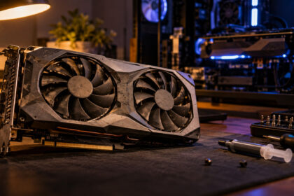 Used graphics card under a desk lamp with dusty fans, an open test bench PC glowing behind it