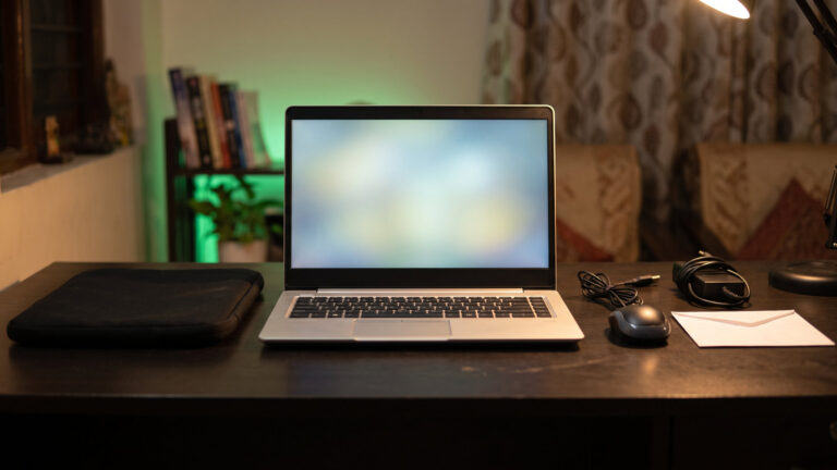 Refurbished silver laptop on a study desk with a laptop sleeve, mouse, charger and a sealed envelope laid out